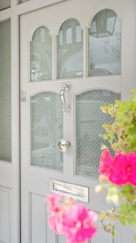a grey front door with chrome hardware and etched glass, behind pink flowers